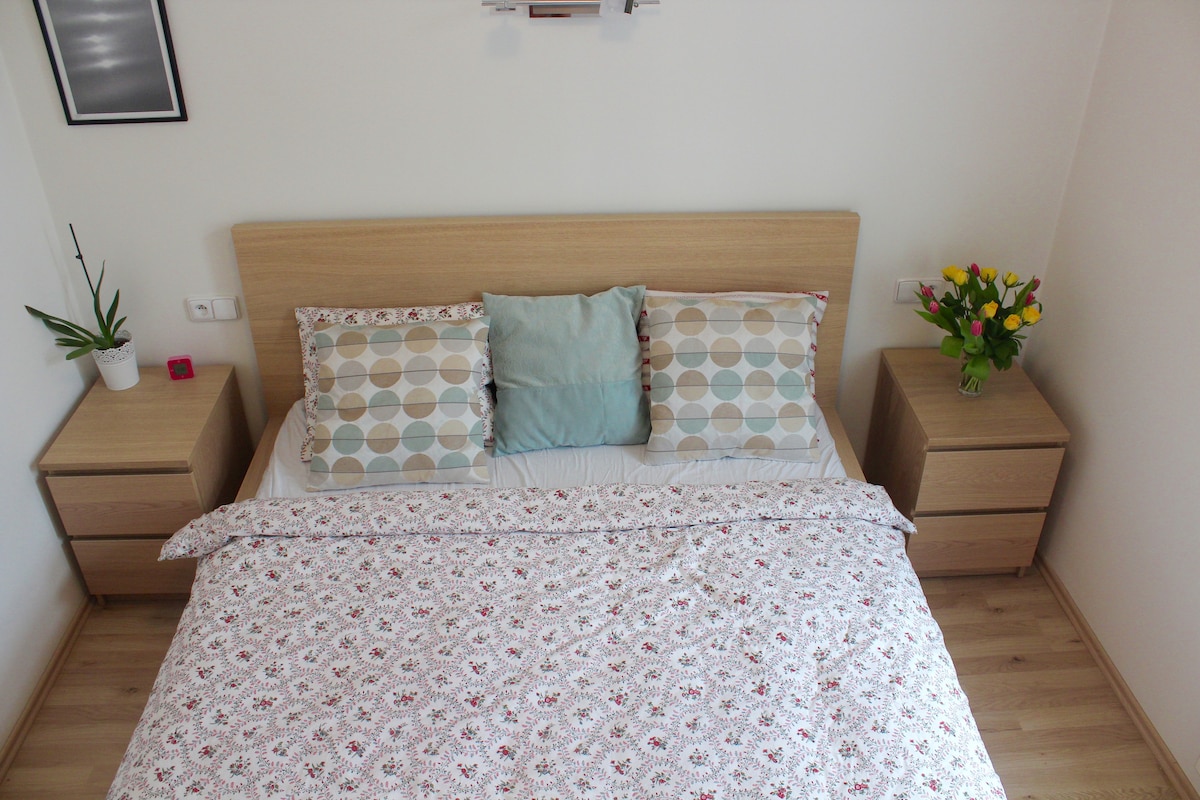 A bright sleeping area features a spacious bed with a floral-patterned duvet and decorative pillows. Two wooden nightstands flank the bed, each with a small lamp. A potted plant and a vibrant bouquet of tulips are visible, adding a touch of color to the room.