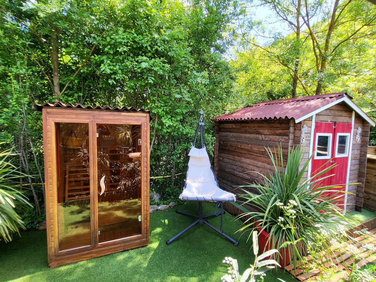 A wooden sauna is positioned beside a charming garden shed, surrounded by lush greenery. A comfortable hanging chair provides a relaxing space, enhancing the outdoor setting. Option for shade is offered by the surrounding plants on the artificial grass.