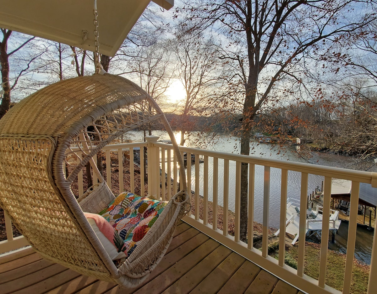 A hanging chair is positioned on a deck, overlooking a tranquil cove. Sunlight reflects gently off the water, while bare trees frame the scene. A wooden dock with boat slips can be seen in the distance, creating a serene lakeside atmosphere.