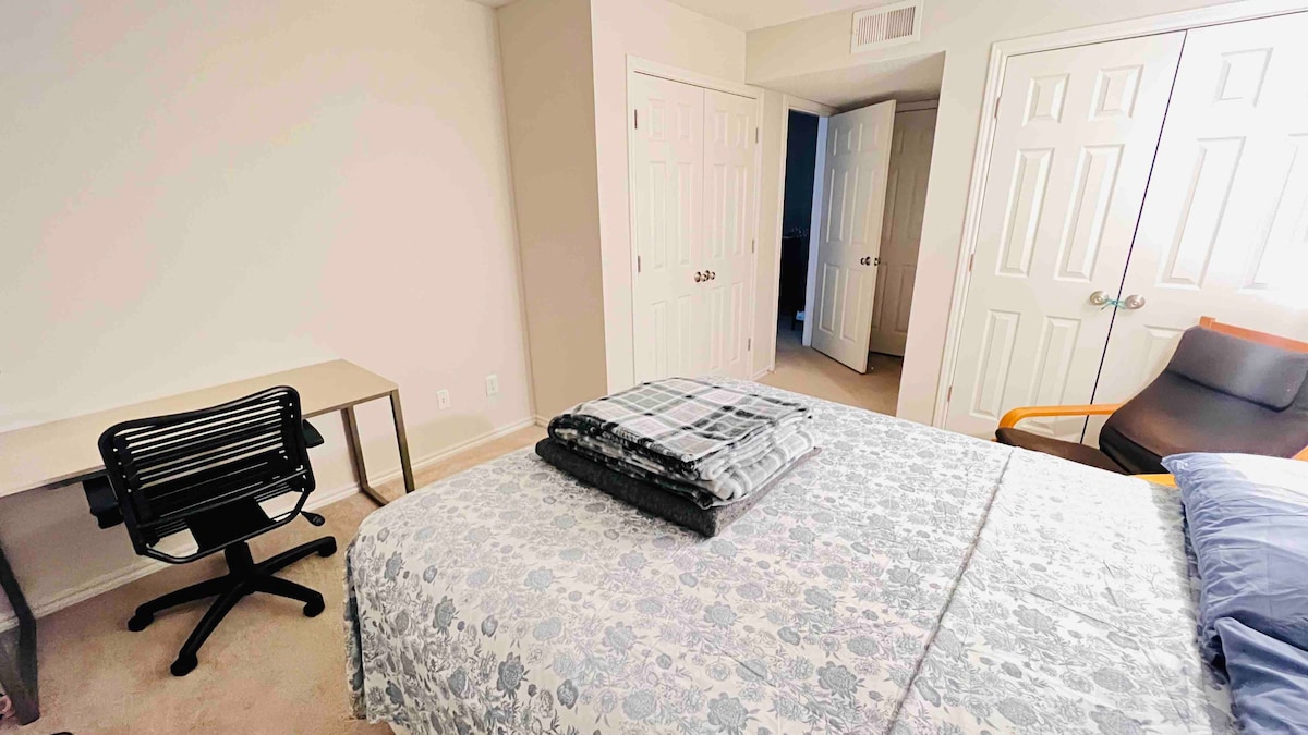 A light-colored bedroom features a spacious bed adorned with a patterned blanket, topped with folded linens. A simple desk and chair are positioned nearby, while light-colored walls enhance the room's brightness. Two doors lead to other areas, contributing to the room's functionality.