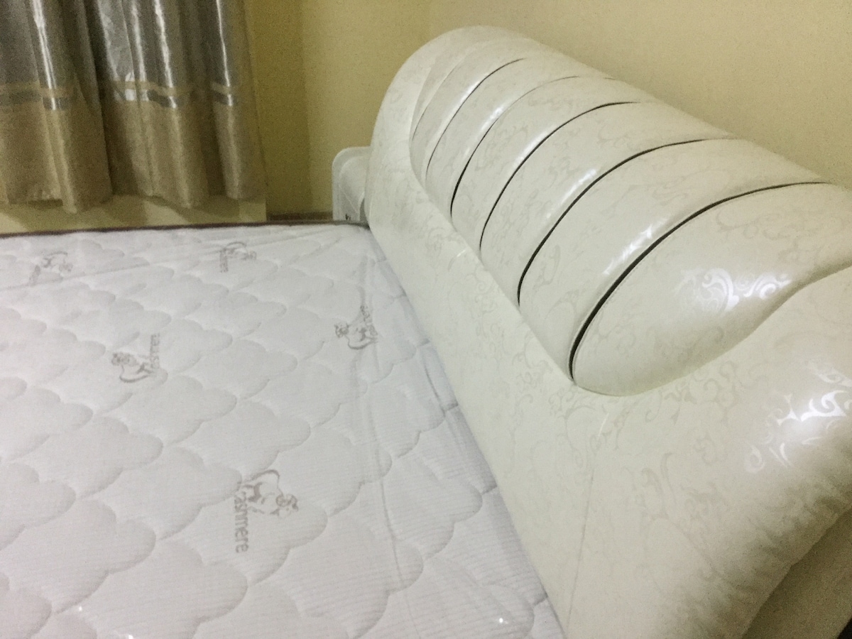 A spacious bed features a decorative headboard with subtle detailing, complemented by a lightly patterned mattress. Nearby, curtains with a soft sheen adorn a window, adding a touch of elegance to the room.