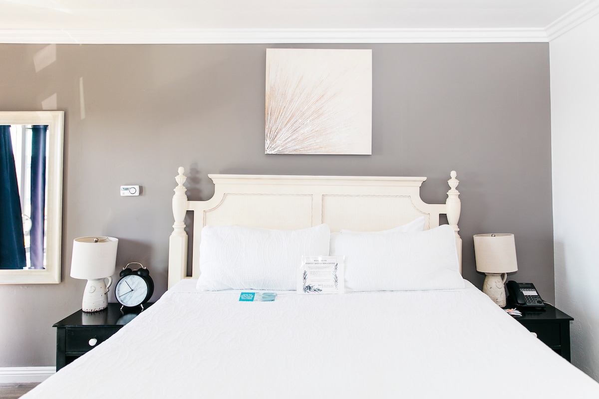 A spacious bed with soft white linens is centered against a neutral wall. Flanking the bed, two sleek black nightstands support lamps and a clock. A subtle piece of wall art adds a touch of decor above the headboard, contributing to the minimalist aesthetic.