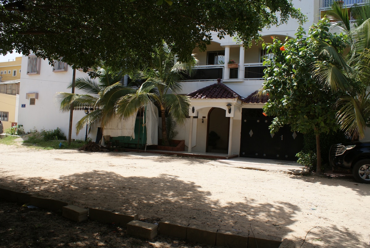 The exterior of a two-story villa is shown, featuring a sandy area with scattered foliage. A covered entrance is visible, complemented by tropical plants. The ground floor includes a garage on the right side, while the upper balcony is adorned with decorative features.