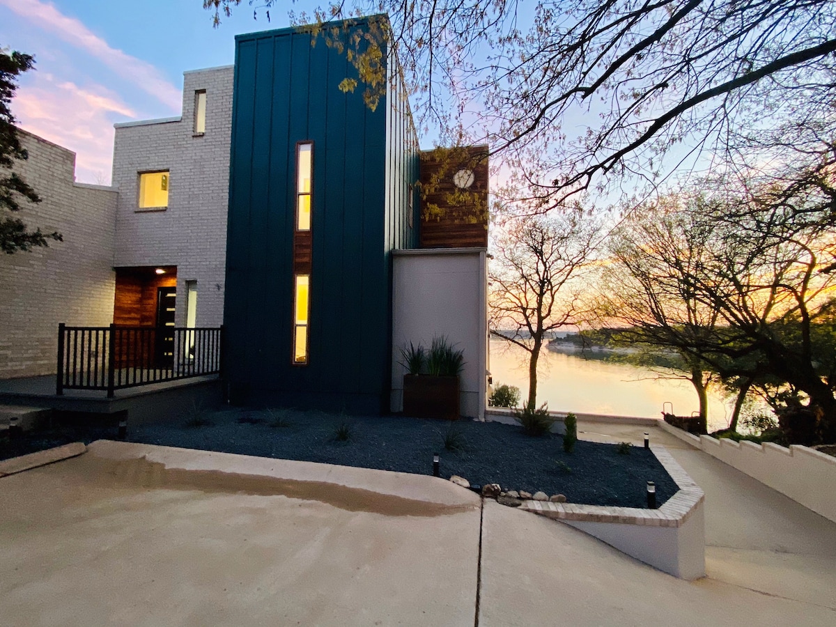 The modern, three-story lake house is surrounded by carefully arranged landscaping and features a distinctive blue exterior. Large windows reflect the soft hues of the sunset, providing a view of the nearby water. A spacious driveway leads up to the structure.