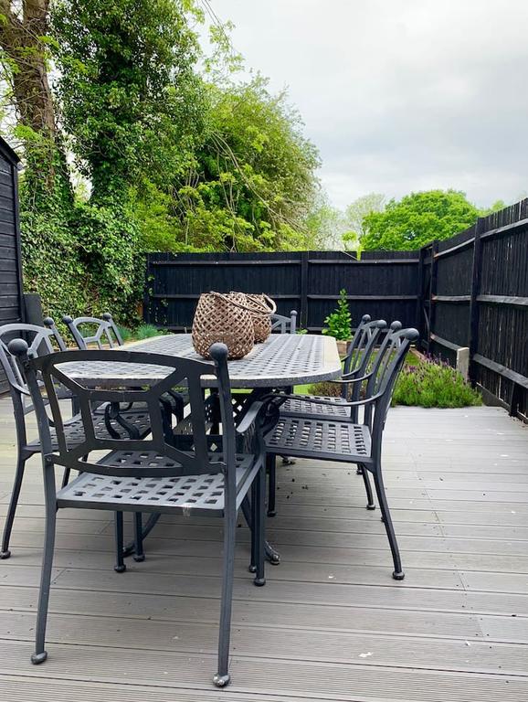 An outdoor dining area features a long, rectangular table surrounded by six metal chairs. A woven basket rests at the center of the table, while greenery and low plants are visible in the background against a dark wooden fence.