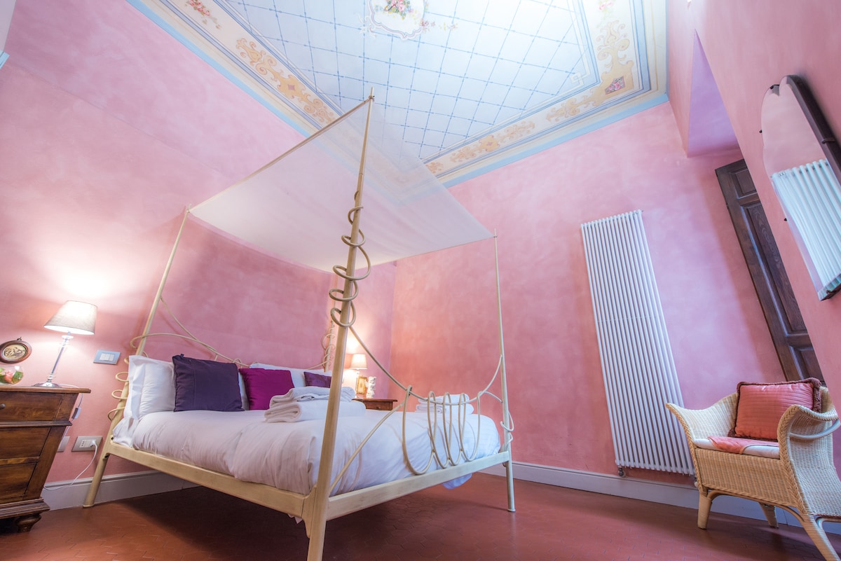 A spacious bedroom features a canopied bed with inviting linens. Soft, pink walls are complemented by a decorated ceiling, adding a touch of elegance. A wooden nightstand and a wicker chair provide additional furnishings, while warm lighting creates a cozy atmosphere.
