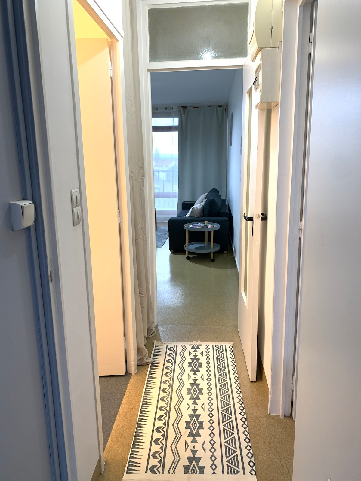 A well-lit hallway is lined with light-colored walls, featuring a decorative rug with geometric patterns. A cozy seating area is visible at the far end, accompanied by a side table. Natural light filters through sheer curtains, creating an inviting path to the living space.