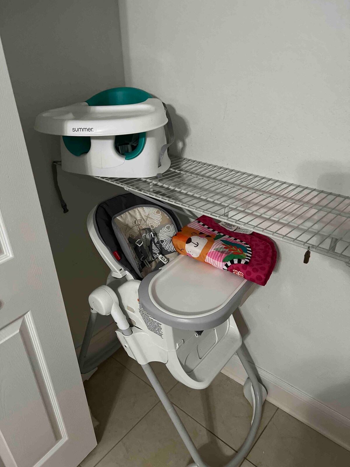 A high chair is positioned next to a wire shelf, which holds a booster seat. A colorful play mat with toys is placed on the chair's tray, providing a convenient space for young children.