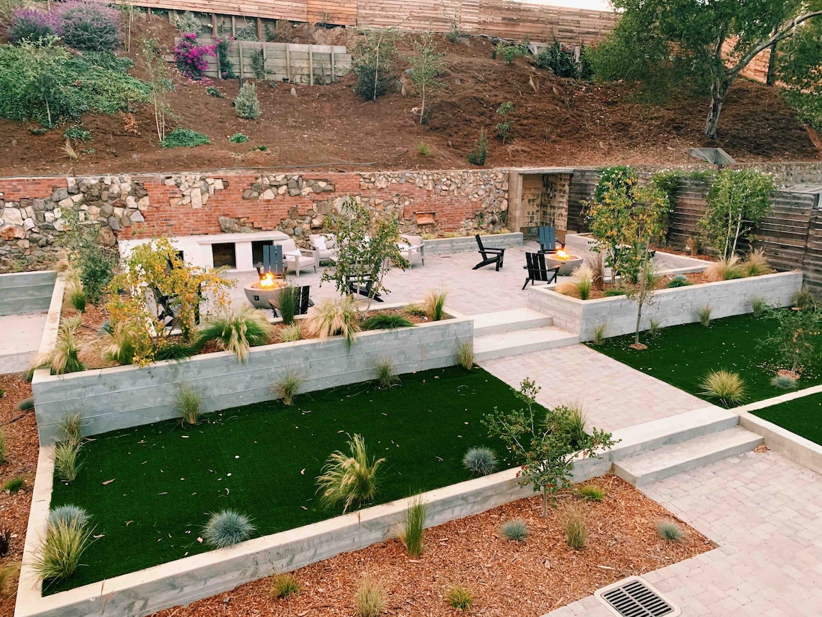 A landscaped outdoor space features multiple stone terraces bordered by greenery and decorative plants. Cozy seating areas with black chairs surround inviting fire pits, providing a warm atmosphere for gatherings. Soft grass-like turf adds a touch of comfort, enhancing the overall serene garden setting.