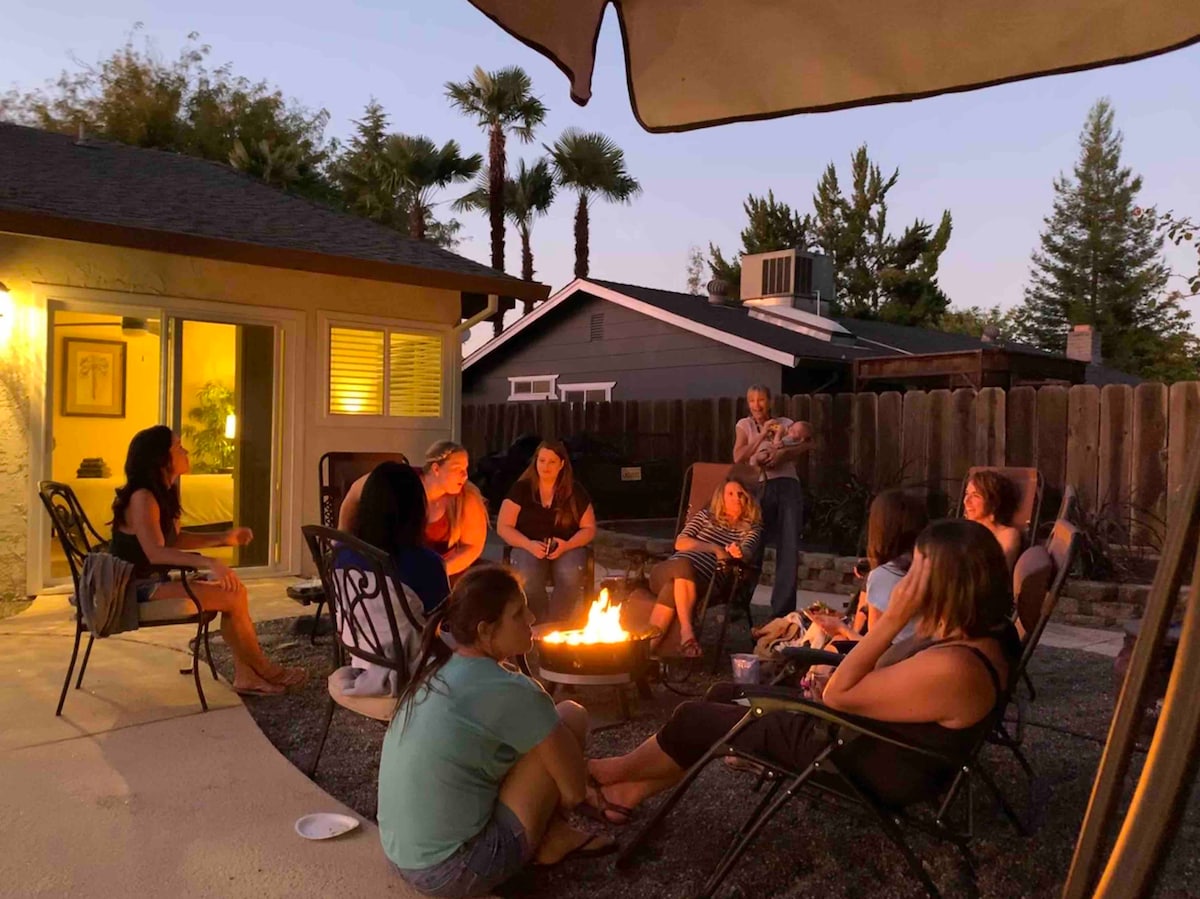 A gathering around a fire pit is depicted, with several guests seated on outdoor chairs, enjoying the evening. Soft lighting from the house and the glow of the fire enhance the relaxed atmosphere, while palm trees and a wooden fence provide a serene backdrop.