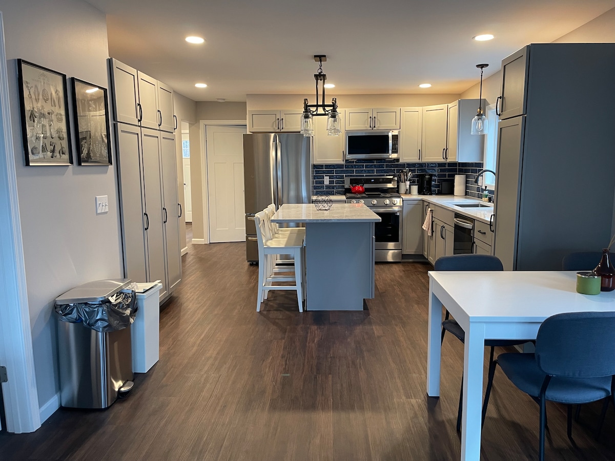 A modern kitchen and dining area features stainless steel appliances and ample cabinetry with a light gray finish. A dining table is set for four, and a white island provides additional seating. Natural light fills the space, highlighting the dark flooring and decorative elements.