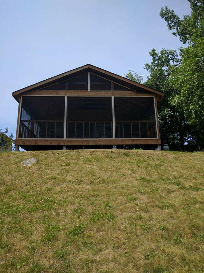 A cottage is depicted on a gentle slope, featuring a large screened-in deck supported by pillars. The structure is framed by several trees, showcasing a natural setting. The deck appears spacious, inviting relaxation with a view over the surrounding grassy area.