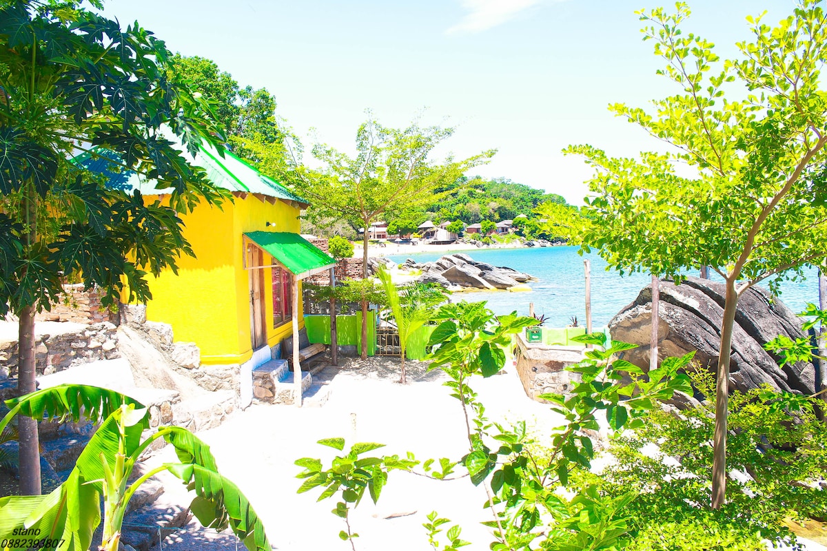 A bright yellow cottage is positioned next to a sandy beach, surrounded by lush green trees. Clear waters are visible nearby, with rocky formations adding natural beauty to the scene. The inviting landscape merges nature with the cozy cottage setting.