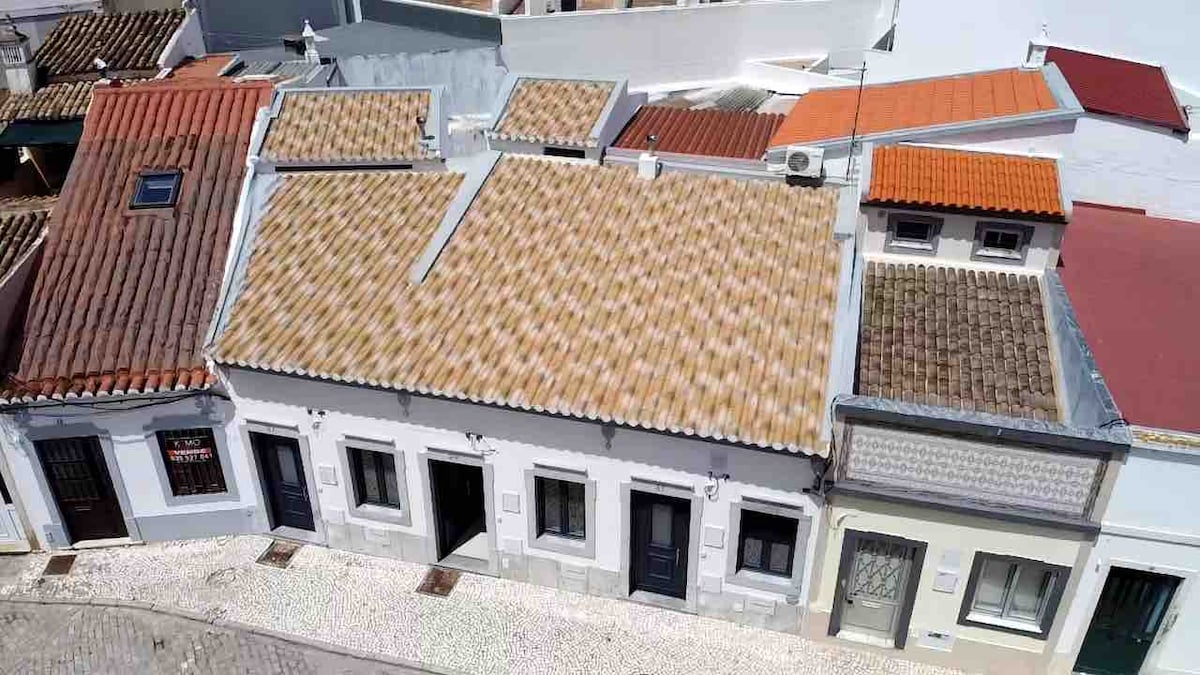 An aerial view captures the charming white exterior of the house, complemented by a terracotta tile roof. Adjacent buildings showcase a mix of traditional and modern architecture, with varied roof colors and textures that enhance the overall urban landscape.