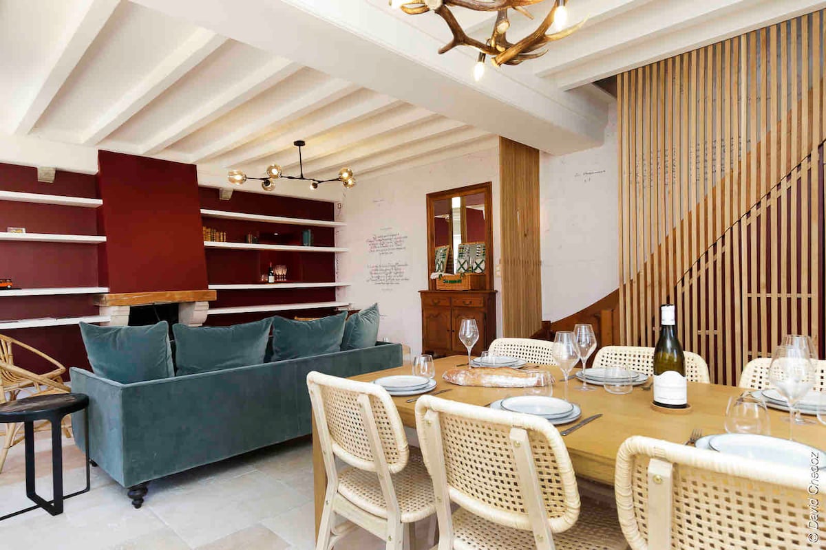 An inviting living area is presented, featuring a plush green sofa and a wooden dining table set with glassware and table settings. Natural light floods the space through the windows, highlighting the elegant mix of textures and colors in the decor.