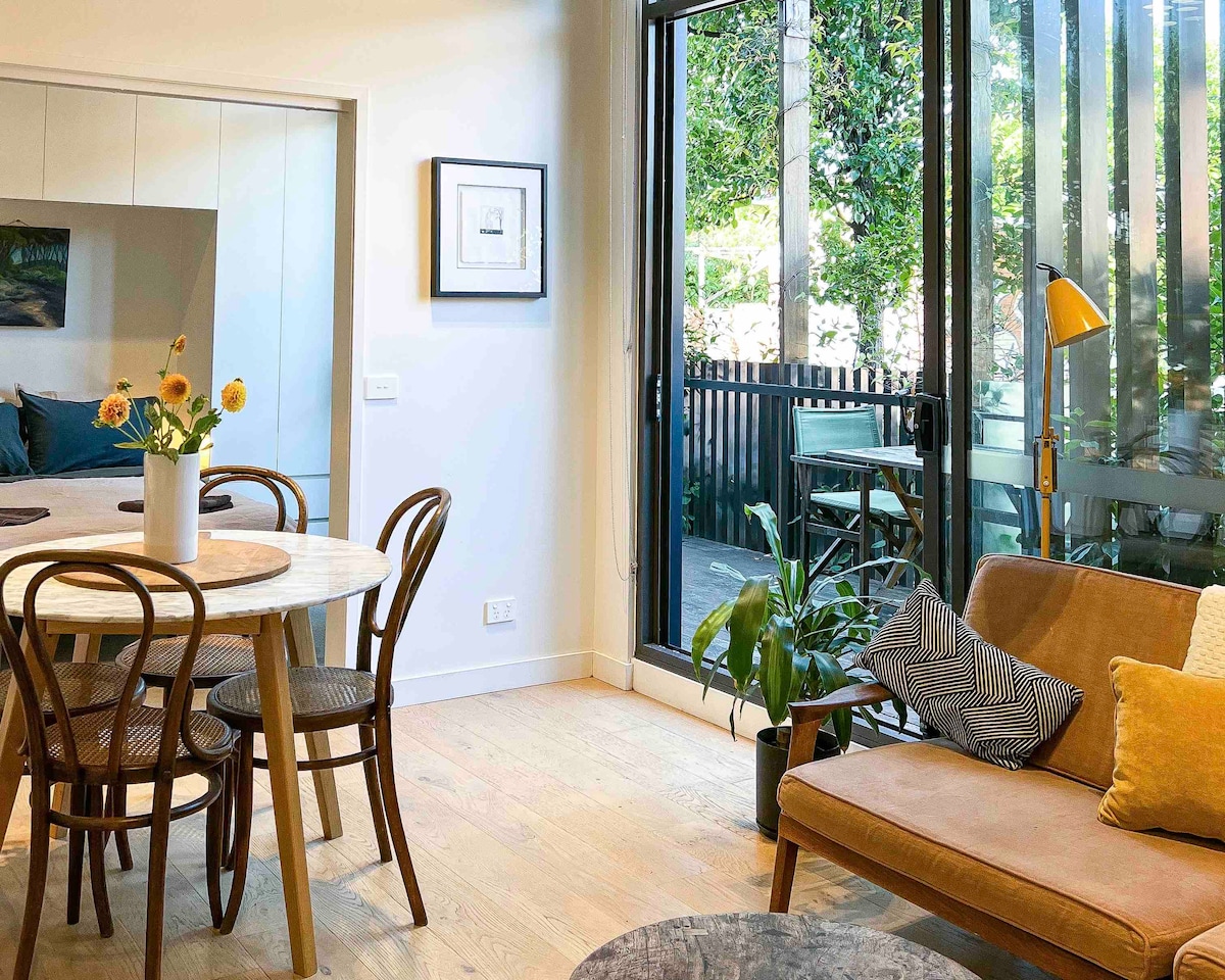 A light-filled living space showcases a round dining table surrounded by wooden chairs, complemented by a cozy brown sofa with patterned cushions. Large glass doors open to a patio, framed by greenery, allowing natural light to enhance the warm wood flooring.