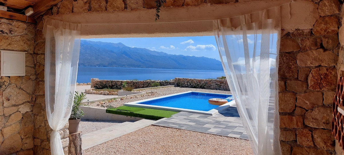 A serene outdoor view is framed by sheer curtains, showcasing a clear blue swimming pool and surrounding lounge area. The distant mountains are visible against the sky, with landscaped stones and greenery enhancing the peaceful setting.