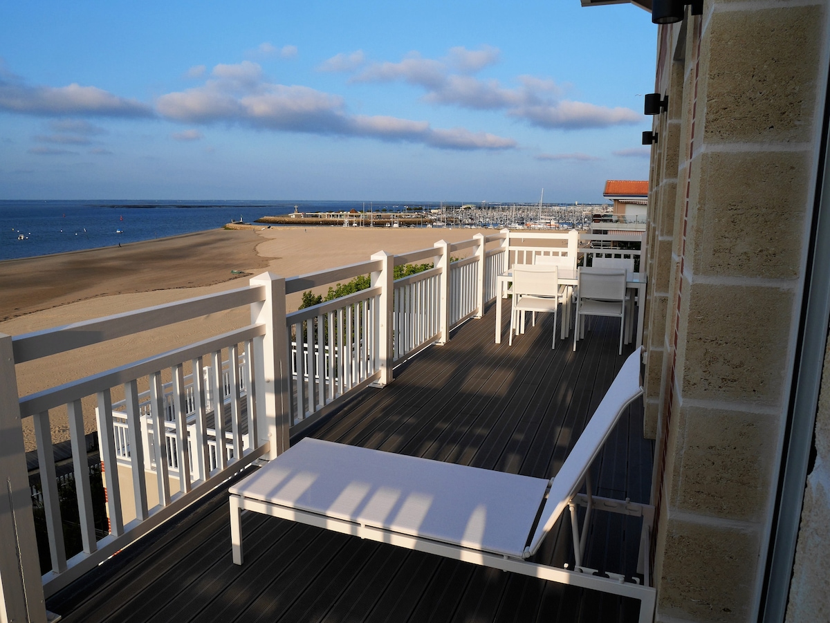 A spacious terrace offers uninterrupted views of the beach and coastline. White lounge chairs and tables are positioned for comfort, while the surrounding railing allows for an open, airy feeling. Gentle waves are visible in the distance, enhancing the serene seaside atmosphere.