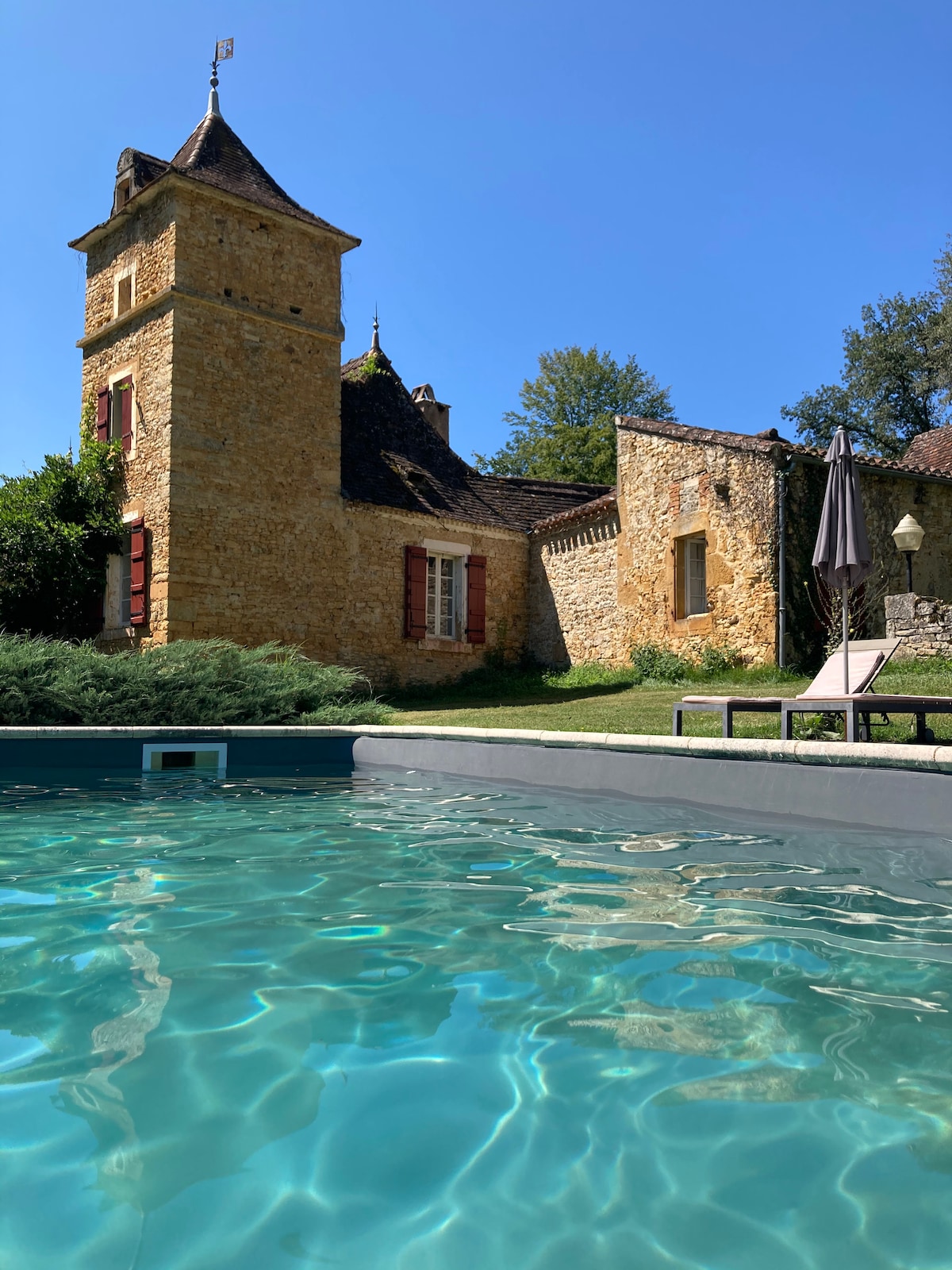 A tranquil outdoor area features a clear swimming pool reflecting sunlight. A rustic stone tower stands nearby, complemented by greenery and a cozy lounge chair, creating a relaxed setting. The building's rustic charm is accentuated by its warm-colored stone and distinctive roof.