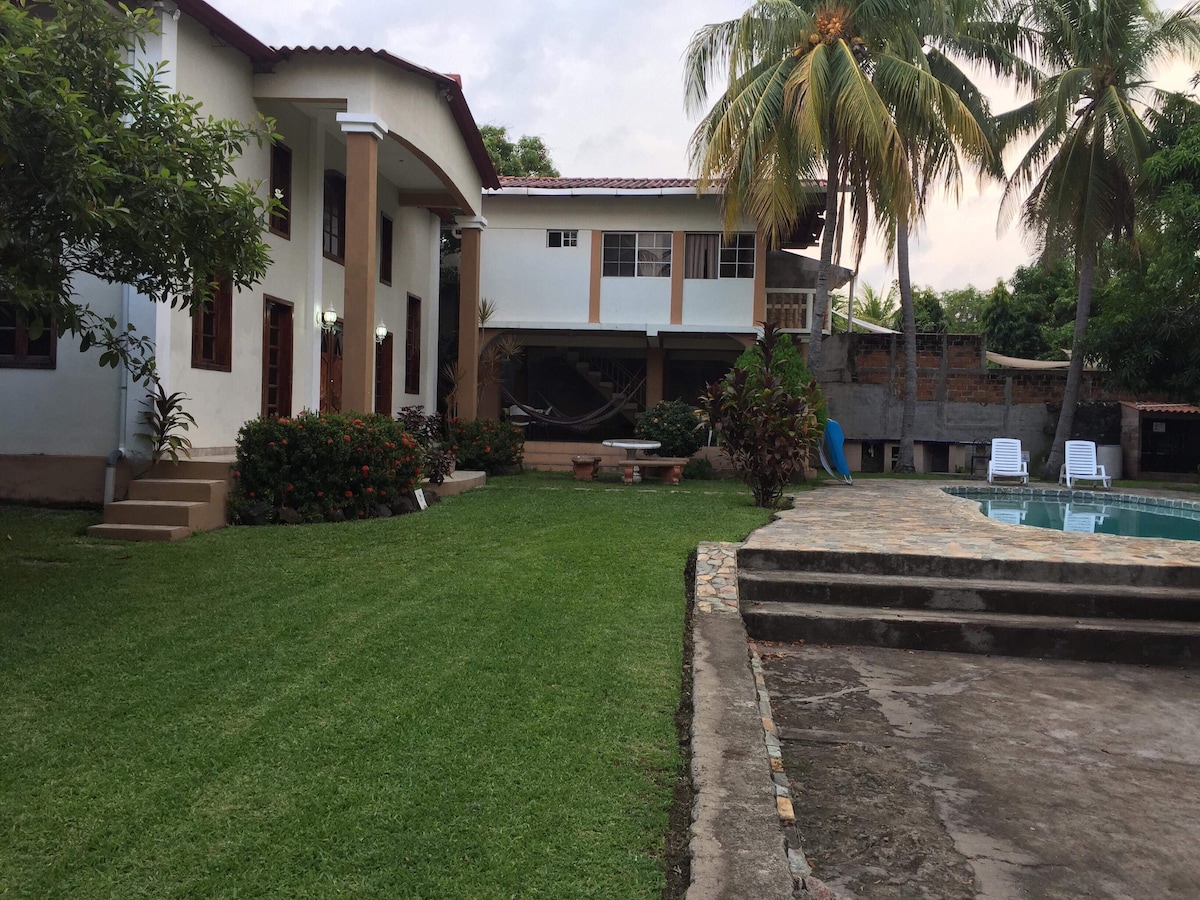 A spacious lawn bordered by well-maintained trees and vibrant shrubs surrounds a two-story home. A clear swimming pool is visible in the background, with sun loungers arranged nearby. A stone pathway leads from the house to the pool area, enhancing outdoor accessibility.