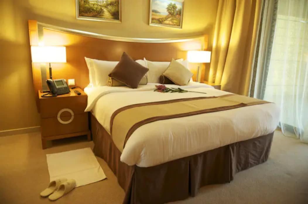 A spacious sleeping area features a large bed with decorative pillows and a subtle runner. Bedside tables with lamps are positioned on either side. A telephone is visible, and a bouquet of flowers is lightly placed on the bed, with plush slippers arranged on the floor.