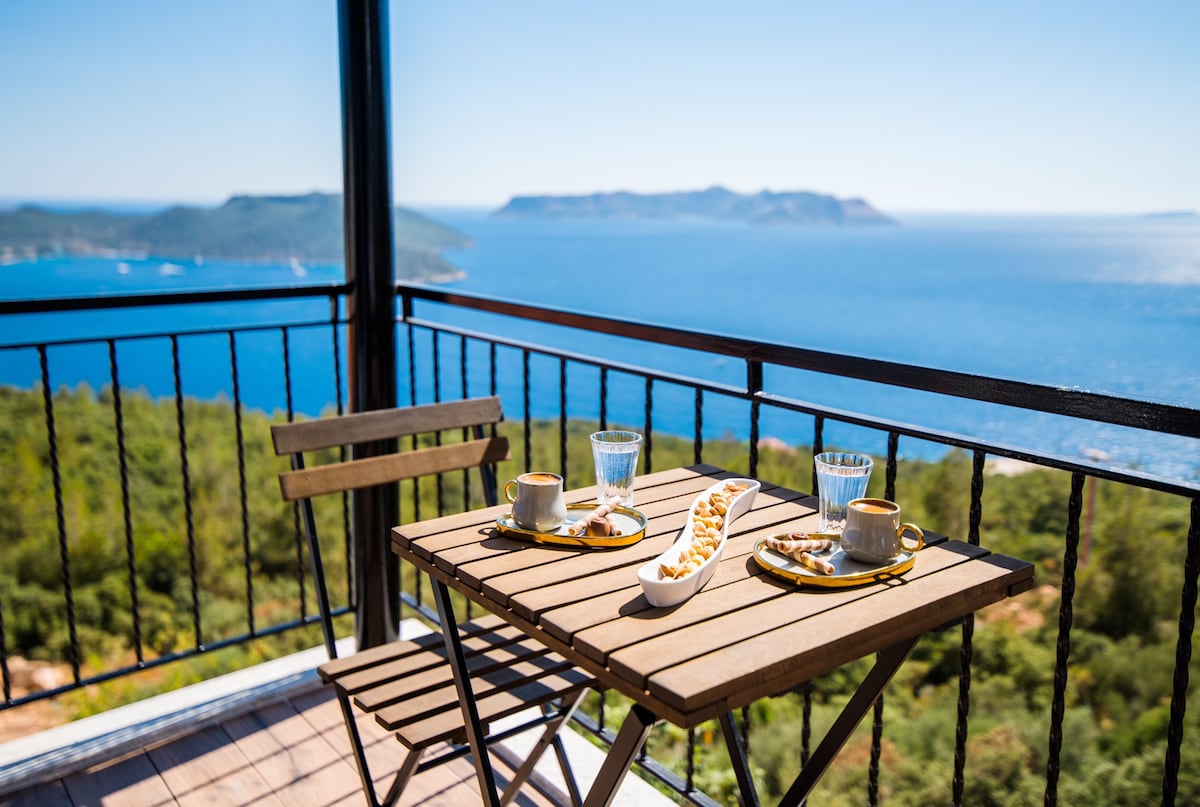 A balcony table set for two features ceramic plates with snacks, accompanied by glasses of water and coffee cups. The backdrop showcases a panoramic view of the sea and distant islands, framed by a black metal railing and lush green hills.