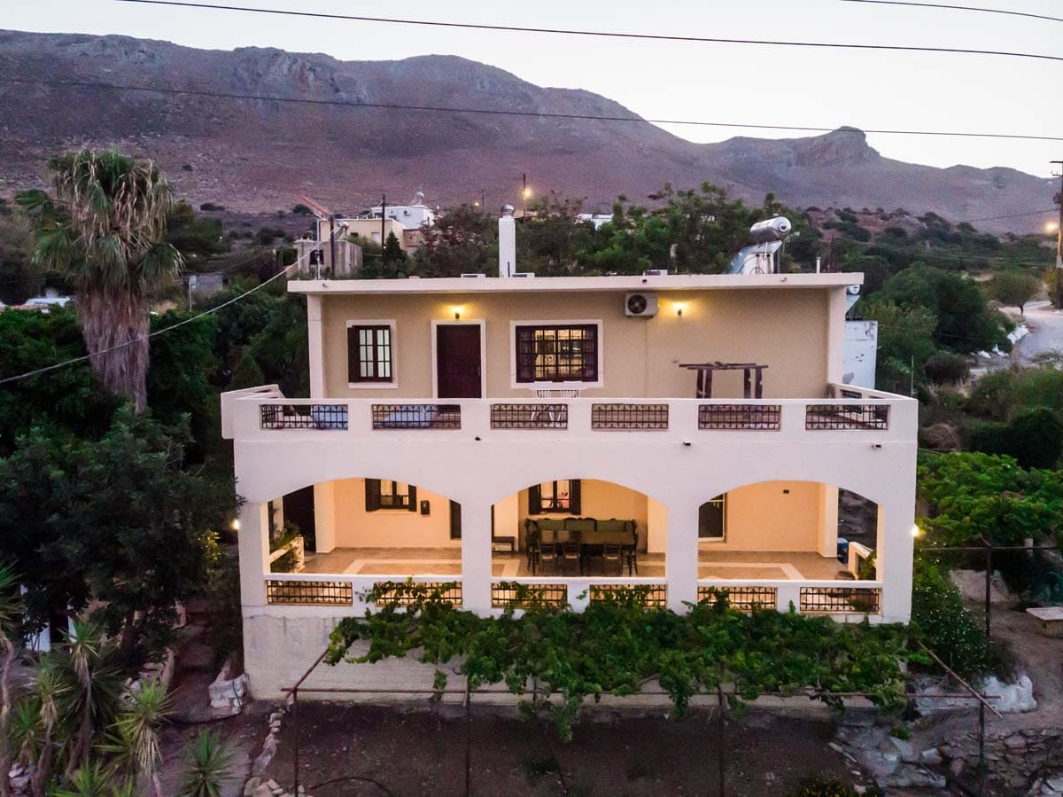 The two-story villa is set against a mountainous backdrop, featuring a wide terrace with arched openings. A dining area can be seen under the terrace, surrounded by greenery. Palm trees and various plants are positioned in the garden area, enhancing the outdoor space.
