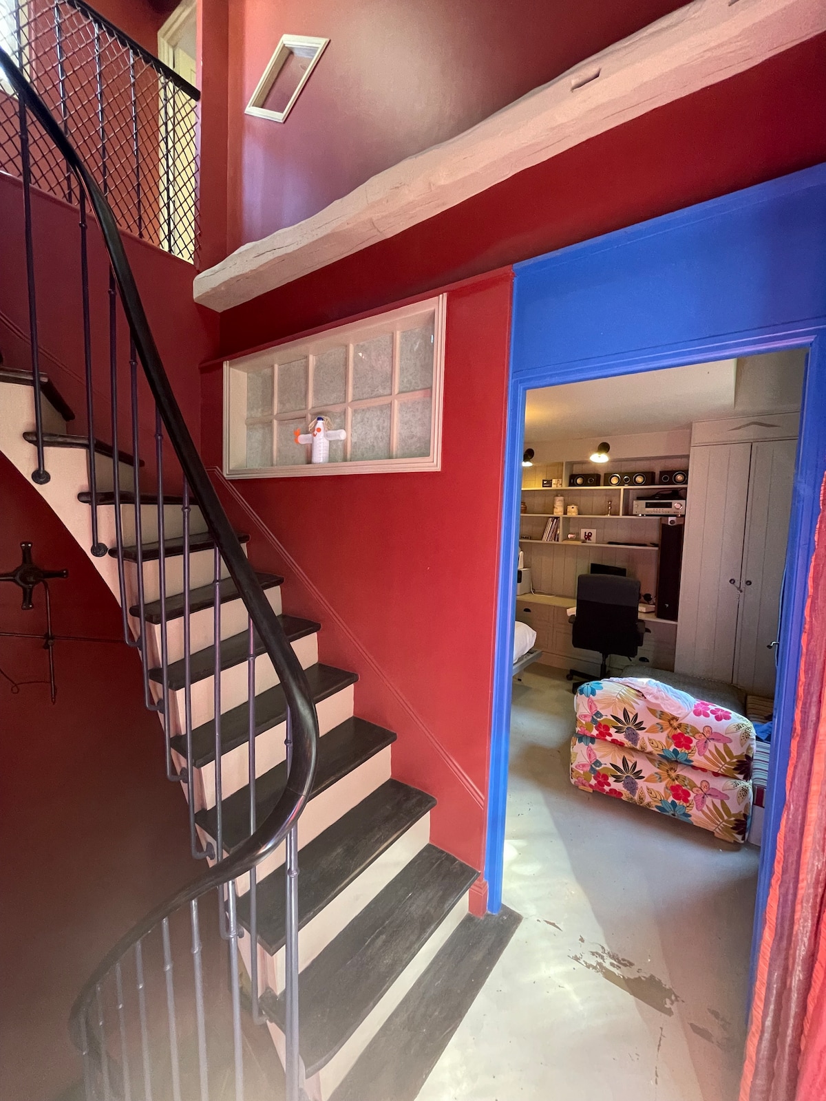 A spiral staircase winds upwards against a deep red wall, framed by rustic wooden beams. A bright blue door leads to a room with shelving and colorful furnishings, inviting a sense of creativity and comfort.