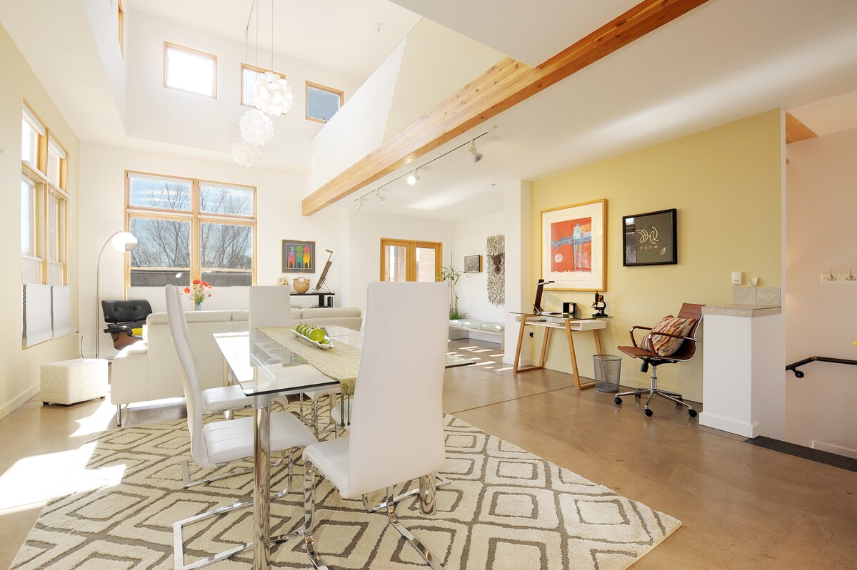 A spacious dining area features a modern glass table surrounded by white chairs, complemented by a patterned area rug. Natural light fills the room through large windows, highlighting artwork on the walls. A comfortable seating area with a chair is visible in the background.