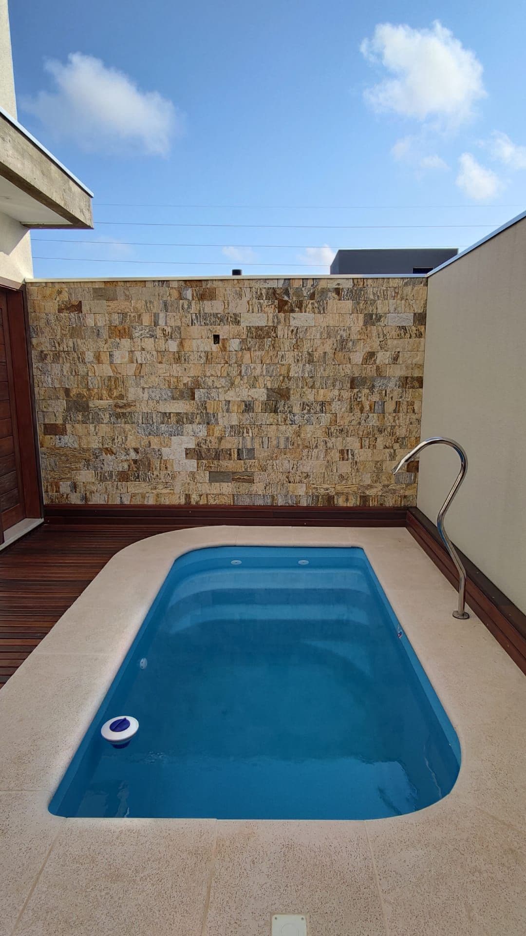 A small swimming pool features clear, inviting water complemented by a smooth, light-colored stone deck. The surrounding walls are decorated with textured stone tiles, while the sky above provides a backdrop of soft, wispy clouds.