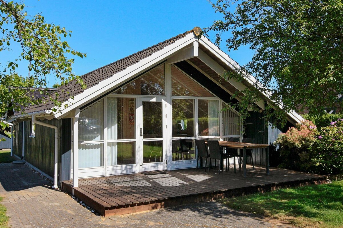 A single-story holiday cottage is set in a peaceful outdoor environment. The property features a wooden deck at the entrance with a table and chairs, surrounded by greenery. Large windows allow natural light to fill the interior, contributing to a welcoming appearance.