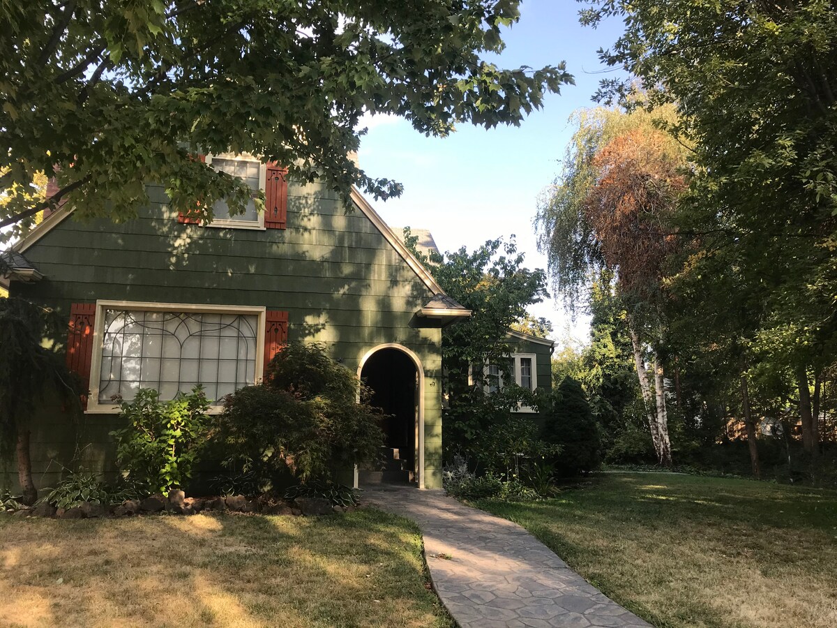 A charming green house with orange shutters is surrounded by trees and shrubs. A stone pathway leads to a welcoming arched entrance, with a well-maintained yard featuring various mature plants and a grassy area.