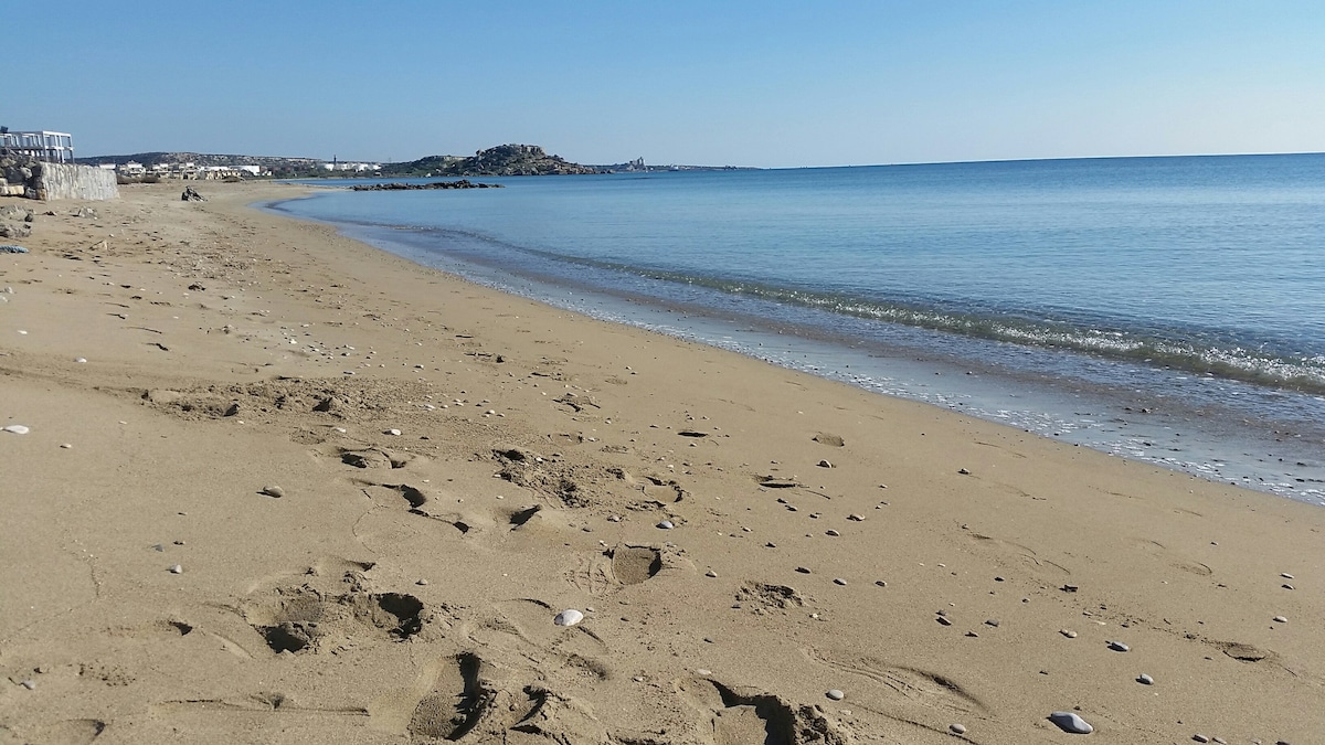 A serene beach scene captures a gently sloping shoreline with soft golden sand. The calm, clear water reflects the bright blue sky. Footprints lead toward the water's edge, where small stones are scattered, adding texture to the sandy landscape.