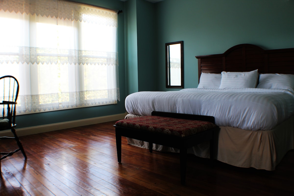 A well-lit room features a spacious bed with white bedding and decorative pillows, positioned against a mint green wall. Natural light filters through large windows adorned with lace curtains. A simple mirror and a wooden chair with a woven seat are positioned nearby, enhancing the serene atmosphere.