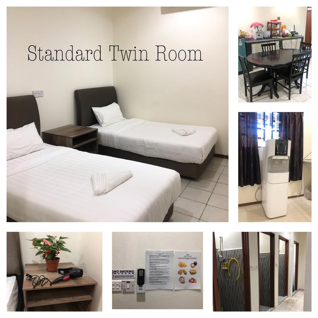 A standard twin room is presented with two spacious beds arranged side by side. A dining table with chairs is visible, along with a kitchenette featuring various appliances. A potted plant sits on a bedside table, while essential amenities are mounted on the wall.