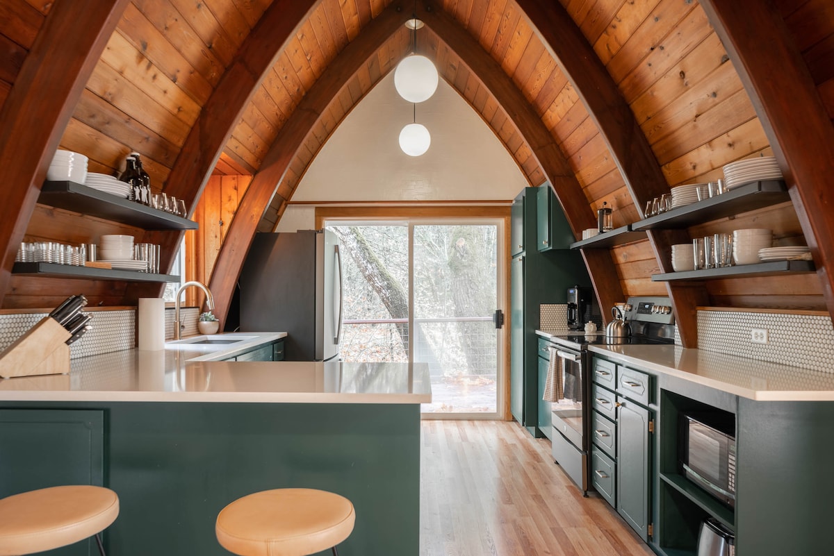 A modern kitchen is showcased within the A-frame cabin, featuring wooden beams and large windows that invite natural light. Sleek countertops and ample cabinetry provide space for cooking. Essential appliances, including a refrigerator and microwave, are visible, alongside neatly arranged dishware and glassware on open shelves.
