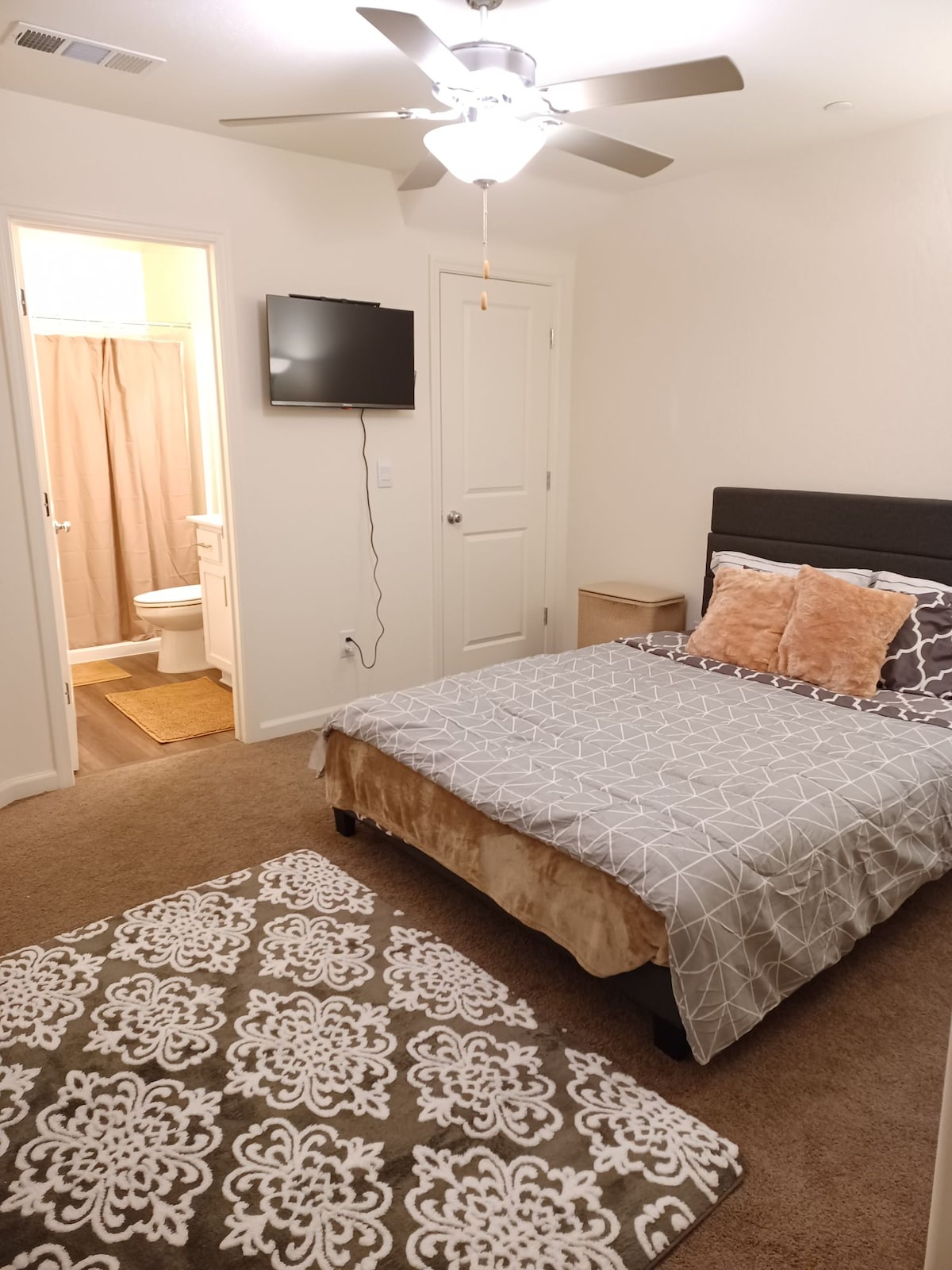 A cozy bedroom features a spacious bed dressed in patterned bedding. A flat-screen television is mounted on the wall, and a door leads to a private bathroom. Soft lighting illuminates the room, complemented by a decorative area rug on the carpeted floor.