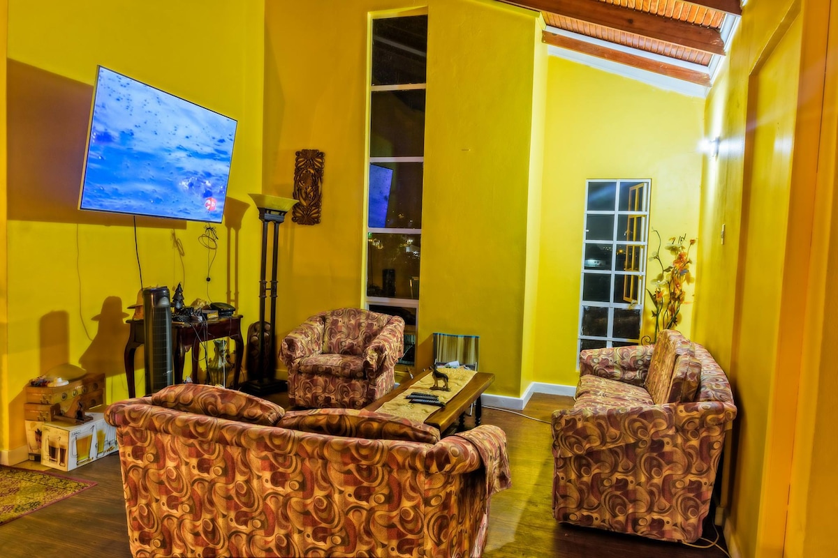 A vibrant living area features a large flat-screen television mounted on a bright yellow wall. Two comfortable sofas, patterned in subtle tones, are arranged around a coffee table. Windows allow natural light, enhancing the spacious feel of the room with a cozy atmosphere.