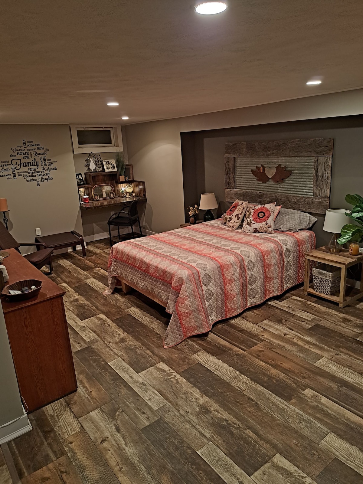 A spacious bedroom is featured with a queen bed adorned in textured bedding. A work desk and chair are positioned against the wall, with soft lighting provided by bedside lamps. Modern decor and wood-like flooring create a warm ambiance throughout the room.