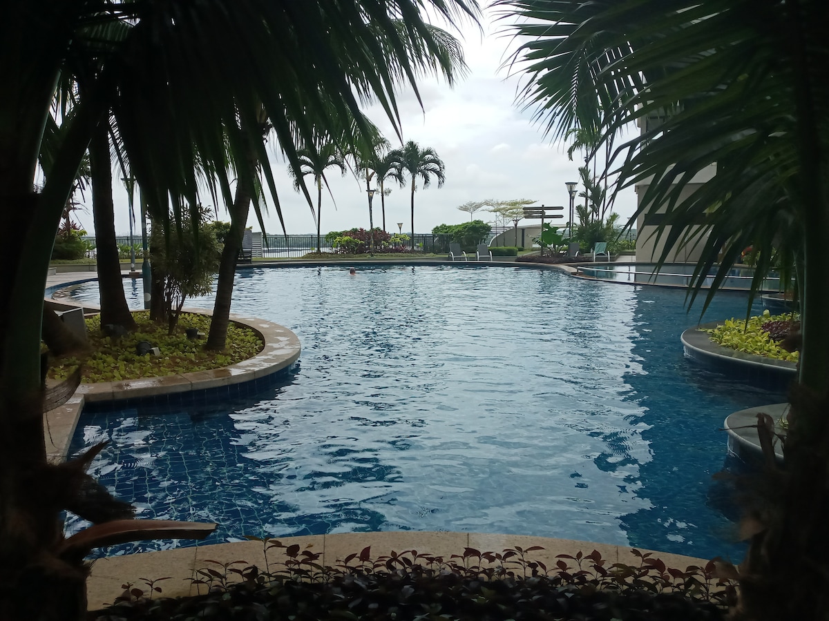 A large swimming pool is framed by lush palm trees, with serene water reflecting the sky. Comfortable lounge areas and green landscaping can be seen around the pool, offering a calm retreat for guests. The beach is visible in the distance.