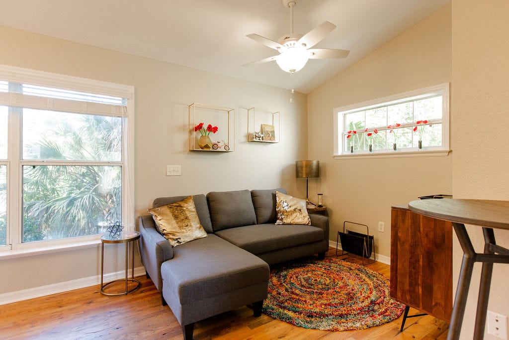 A comfortable living area features a sectional sofa with decorative cushions, complemented by a colorful rug. Natural light enters through two windows, providing a view of greenery outside. A small table and lamp complete the space, creating an inviting atmosphere.