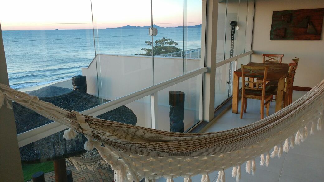 A balcony offers a serene view of the ocean, with a hammock gently swaying and a dining table visible nearby. Large glass panels provide ample light and an unobstructed perspective of the coastline, highlighting the tranquil atmosphere of the space.
