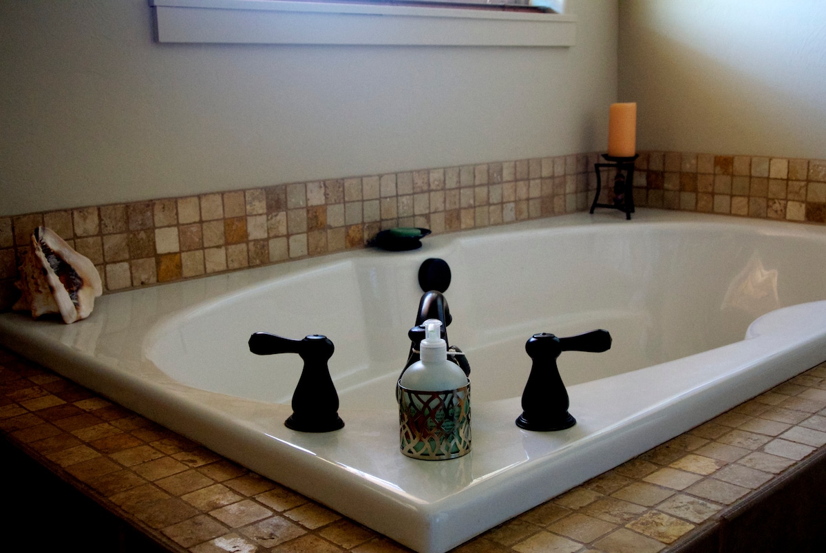 A corner jetted bathtub is featured, surrounded by neutral-toned tile. Two black faucets are visible, along with a bottle of lotion and a candle on a stand nearby. A decorative shell rests on the ledge, adding a subtle coastal touch.