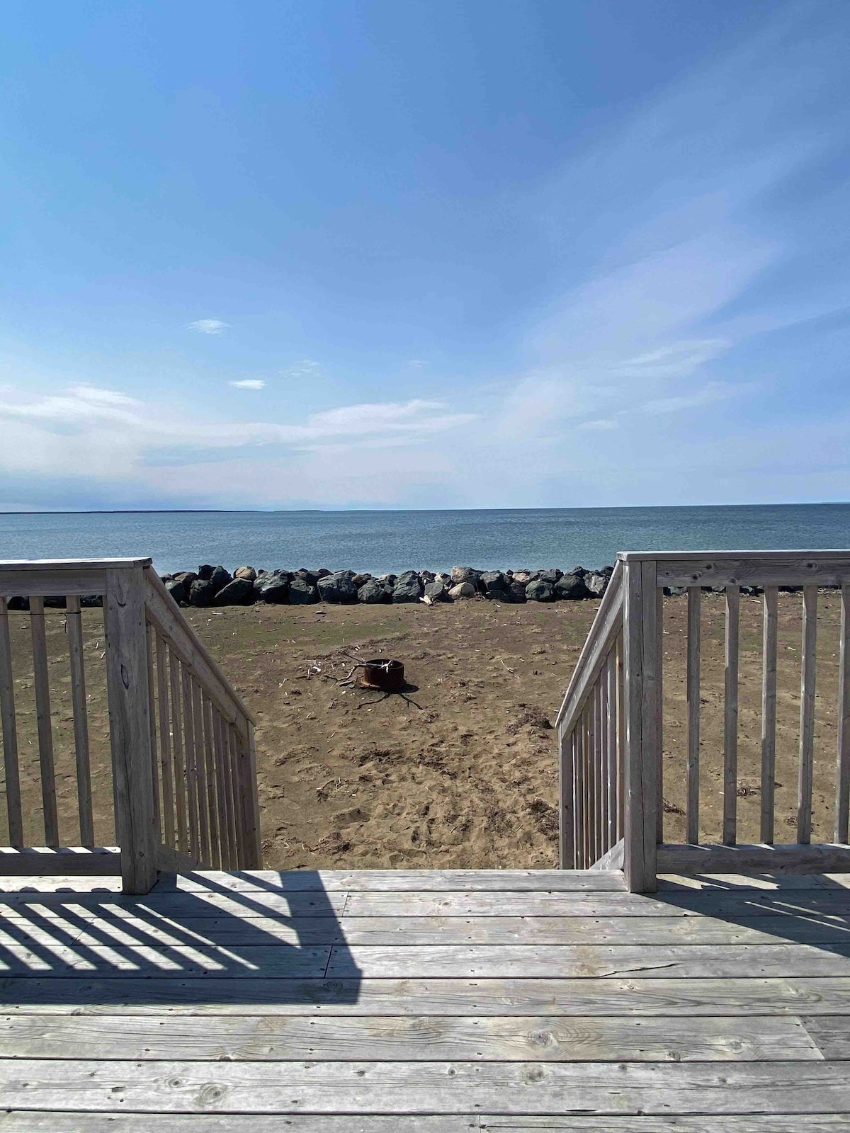 A wooden staircase leads down from a deck to a sandy beach, revealing a calm seascape. Rocks border the shoreline, and gentle waves lap at the shore under a clear blue sky.