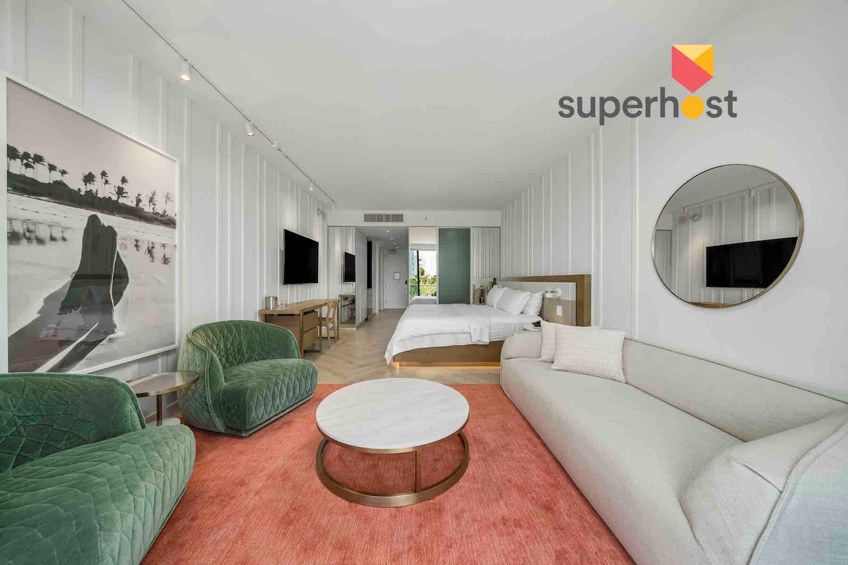The stylish studio features a spacious seating area with green upholstery, complemented by a round coffee table on a soft rug. Natural light floods the room through large windows, creating a bright atmosphere. A well-appointed bed and modern artwork enhance the elegant decor.