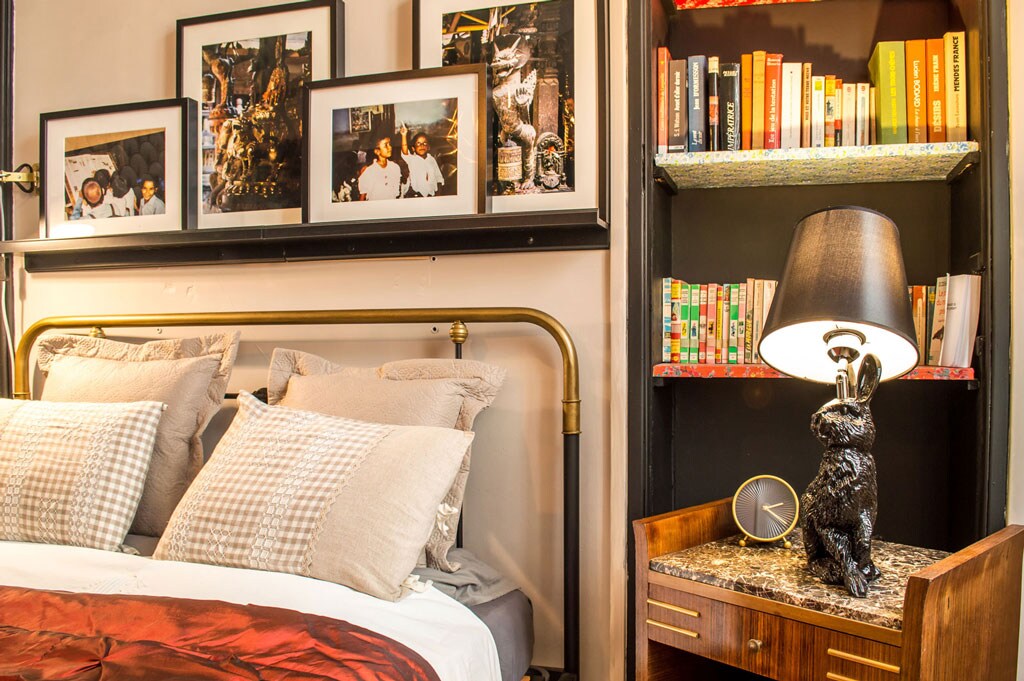 A cozy sleeping area features a brass bed with neatly arranged, patterned pillows. Above the bed, framed photographs add character to the space. A side table, adorned with a decorative lamp, holds a rabbit sculpture and a clock. A bookshelf displays colorful books in a pleasing arrangement.