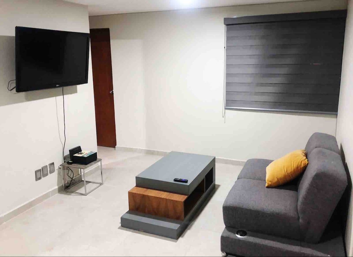 A cozy living area features a gray sectional sofa accompanied by a yellow accent pillow. A multi-tiered coffee table is positioned in front, and a wall-mounted television is visible. A window with dark roller shades allows for adjustable light control.