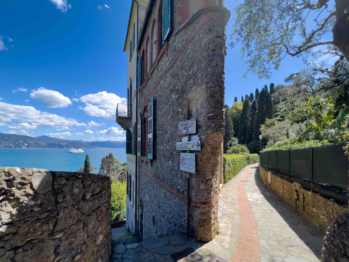 The pathway winds alongside a stone wall, leading to the tower entrance. Lush greenery is visible along the route, with a stunning view of the coastline and sea in the background. The clear sky enhances the serene atmosphere of the area.