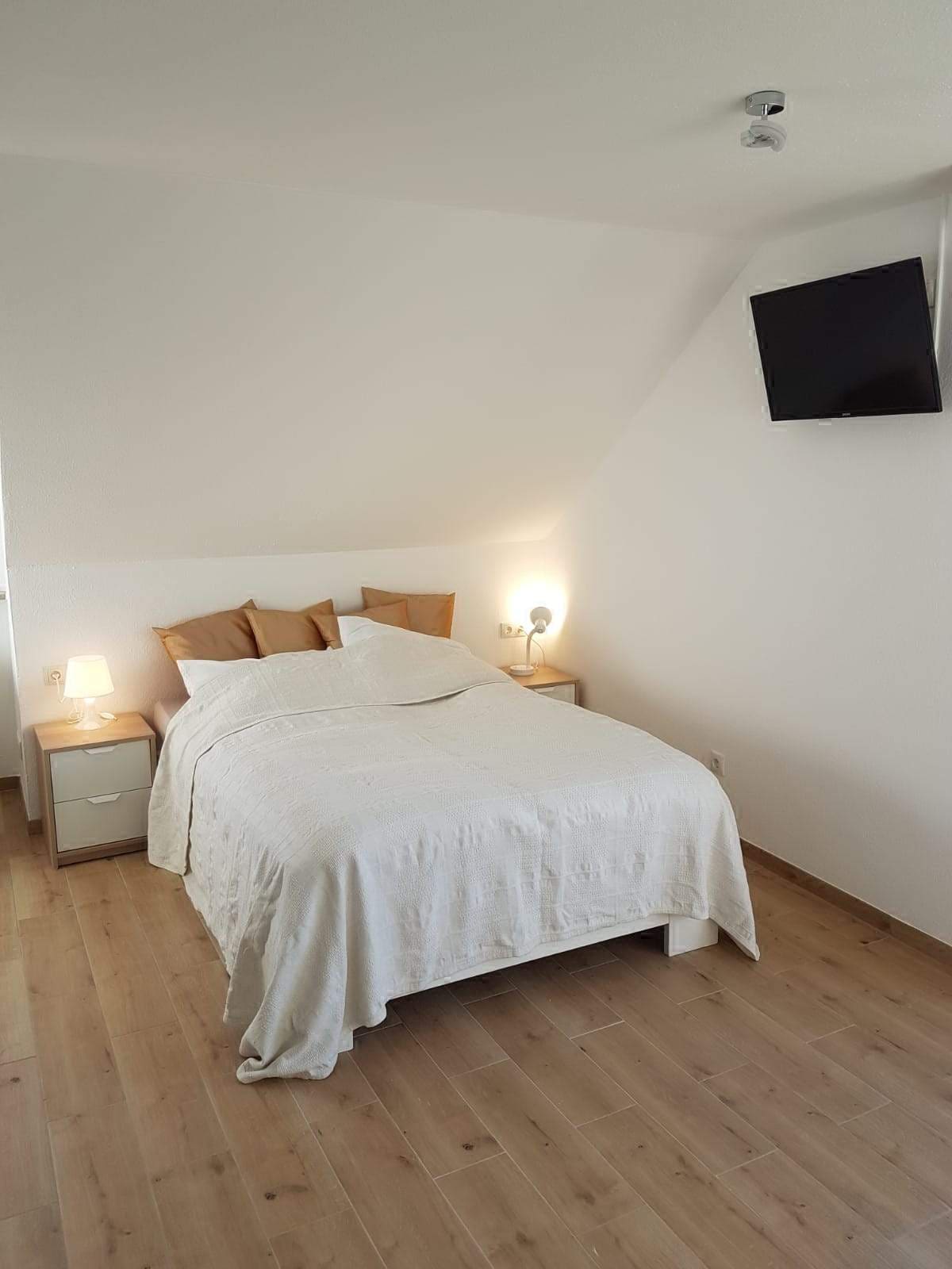 A bright bedroom is presented with a spacious bed dressed in light bedding, flanked by bedside tables with lamps. A wall-mounted television is visible, and soft natural light enters through a nearby window, enhancing the room's calm and minimalist design.