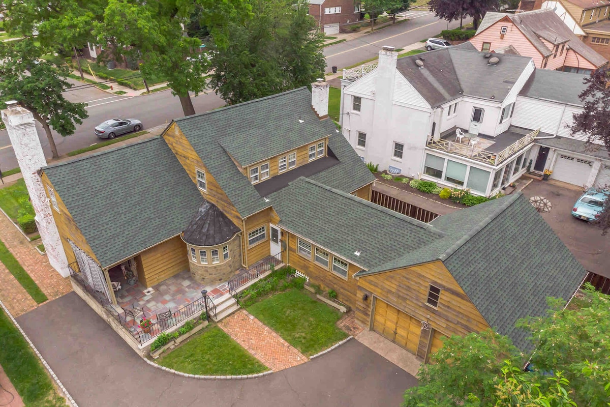 An aerial view presents a spacious house with a unique architectural design, featuring a combination of stone and wood exteriors. The surrounding area includes a neatly landscaped yard and nearby residential houses, highlighting the tranquil neighborhood.